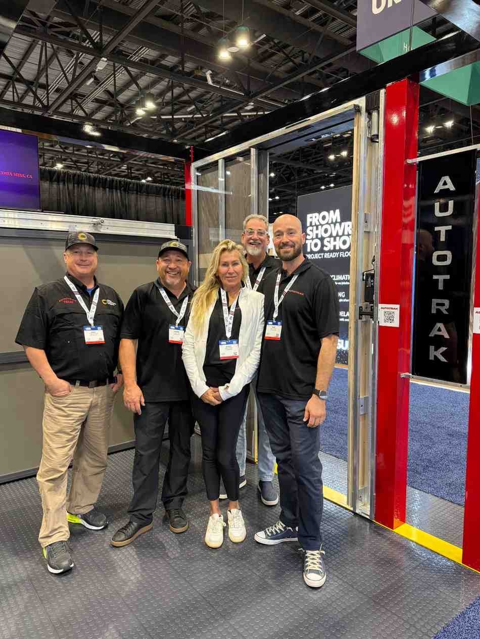 Five people, three men and two women, smiling at a trade show booth that says 'AUTOTRAK' and 'FROM SHOWR TO SHO'.