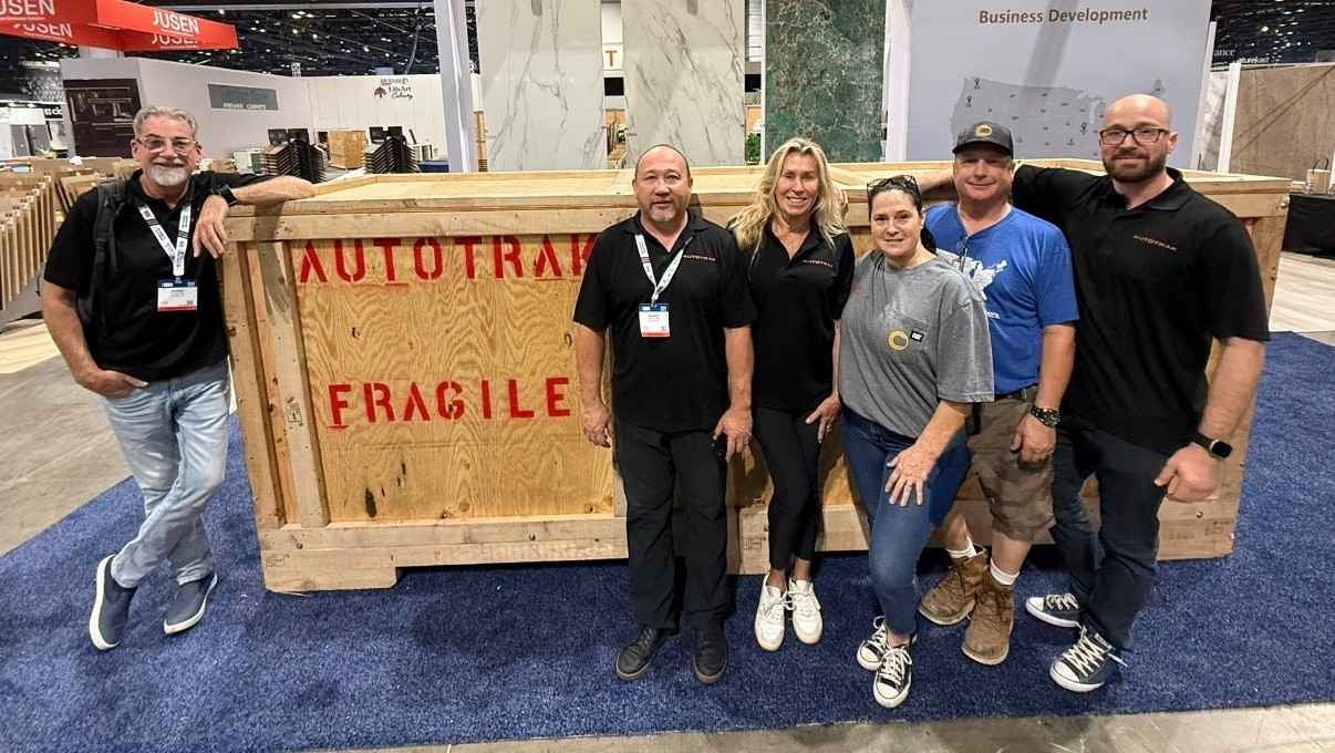 Six people, four men and two women, pose smiling by a large wooden crate marked 'AUTOTRAK FRAGILE.'